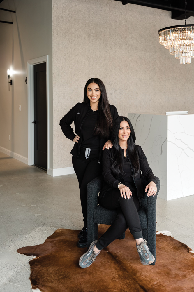 Two women posing in the lobby of their salon. One of them, Amanda Afr, is standing behind a woman who's sat down in a chair with her legs crossed, Asiel Elian.